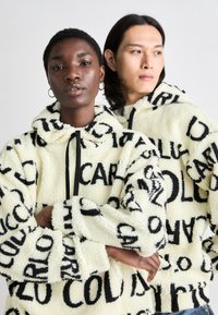To modeller bærer oversized, gule fleece-hoodies med sorte tekstmønstre. Fleecen har en blød tekstur og snorelukninger ved halsen.