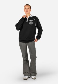 Μαύρο φούτερ με λευκό γιακά και λογότυπο στη στήθος. Συνδυάζεται με γκρίζα κολακευτικά τζιν και αθλητικά παπούτσια. Λείο υλικό.