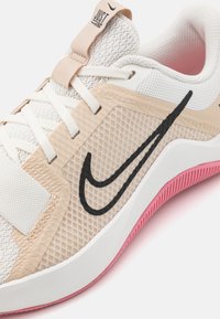 Zapatilla deportiva de color beige claro con parte superior de malla, cordones blancos, logotipo de Nike en negro, suela de goma rosa y detalles texturizados en el lateral.