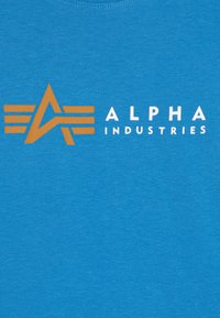 Mėlyna medvilninė marškinėliai su grafinėmis logotipais, kuriose yra oranžinė žvaigždė ir linijos, taip pat baltiems tekstu "ALPHA INDUSTRIES".