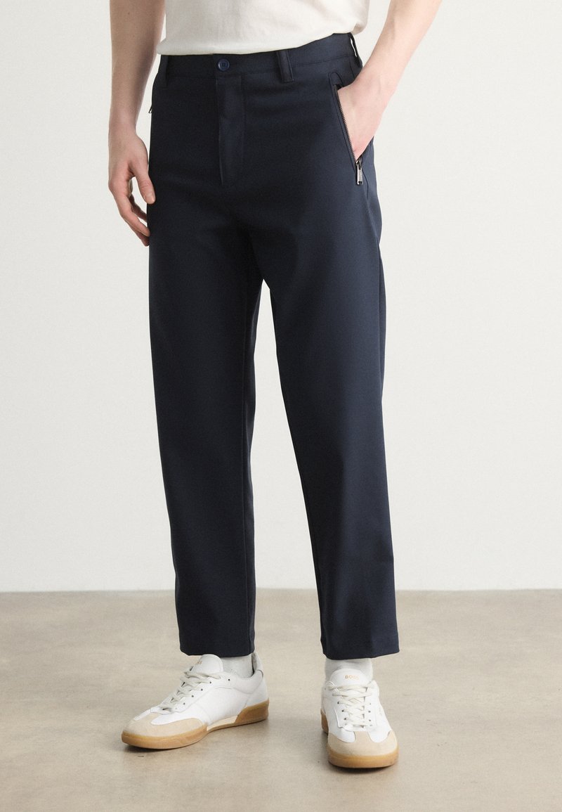 Ναυτικές παντελόνες με χαλαρή εφαρμογή, που διαθέτουν τσέπη με φερμουάρ, επίπεδο μπροστινό μέρος και φάσες στον αστράγαλο, σε συνδυασμό με λευκά αθλητι�κά παπούτσια και κάλτσες μέχρι τον αστράγαλο.