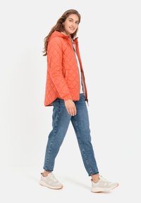 Gequilte oranje jas met capuchon, voorzien van een ritssluiting en twee zijzakken. Gecombineerd met lichtblauwe jeans en grijze sneakers.