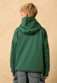 Felpa verde con finitura texturizzata, dotata di un ampio cappuccio e maniche lunghe, abbinata a pantaloni corti in denim blu con tasche a toppa.