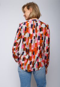 Kolorowa bluzka z długim rękawem z miękkiego, gładkiego materiału z geometrycznym wzorem z prostokątów w kolorze czerwonym, różowym, brązowym i niebieskim. Widoczny detal w postaci zakładki na plecach.
