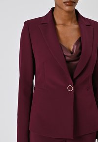 Bordeaux blazer met een getailleerde pasvorm, geknipt revers en een ronde metalen knop. Gladde, gestructureerde stof met een subtiele glans.
