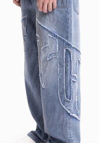 Svetlo modre raztrgane jeans hlače z velikimi srkanimi izrezki, ohlapen kroj in izpran videz. Poudarjeni šivi so jasno vidni.