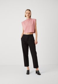 Roze cropped top met brede schouderuitsnijdingen, gecombineerd met zwarte tailored broeken. Model draagt zwarte hoge hakken. Effen achtergrond.