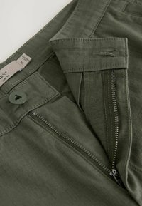 Pantalon slim vert avec fermeture éclair partiellement ouverte, bouton fermé, et détails visibles de la texture du tissu et des coutures.