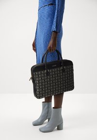 Sac fourre-tout noir et à motifs avec des poignées doubles, doté d'une fermeture éclair. Porté avec une robe bleue à côtes et des bottines grises.