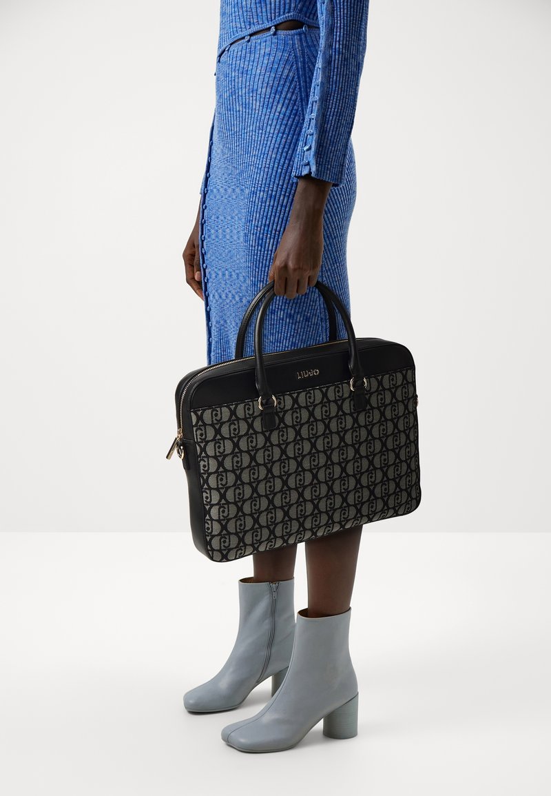 Sac fourre-tout noir et à motifs avec des poignées doubles, doté d'une fermeture éclair. Porté avec une robe bleue à côtes et des bottines grises.