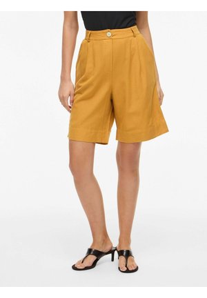 Vrouw draagt mosterdgele high-waist knielange shorts met knoop aan de voorkant, zwarte top en zwarte sandalen met hak en teenring.