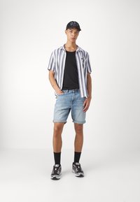 Zilos un baltos svītrains īsām piedurknēm krekls, melna t-krekls, džinsa šorti, melnas zeķes un melnā/baltā sporta apavi, stāv uz viendabīga fona.