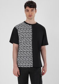 Schwarzes T-Shirt mit einem geometrischen weißen Muster auf der linken Seite. Kurze Ärmel und runder Hals. Baumwollstoff, lockere Passform.
