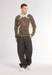 Sweatshirt verde e crema con scollo a V, caratterizzato da un design grafico arancione. Abbinato a jeans scuri, larghi e scarpe da ginnastica bianche.