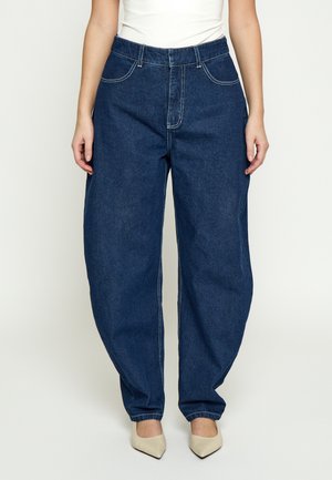 Person iført løstsiddende mørkeblå denimjeans og beige spidse sko, med hænder afslappet ved siden.