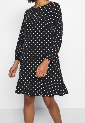 Frau trägt ein schwarzes, langärmeliges Kleid mit weißen Punkten und einem gerafften Saum, steht vor einem schlichten weißen Hintergrund.