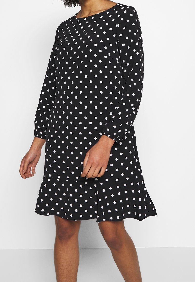Femme portant une robe noire à manches longues avec des pois blancs et un ourlet à volants, debout devant un fond blanc uni.