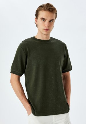 Camiseta de manga corta verde oscuro con cuello redondo, que presenta un corte relajado y un acabado texturizado distintivo. Estilo casual con detalles de puños acanalados.