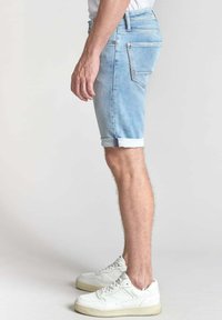 Pantalones cortos de denim azul claro con puños enrollados, diseño de cinco bolsillos y un bolsillo trasero con un parche de color. Llevados con zapatillas blancas.
