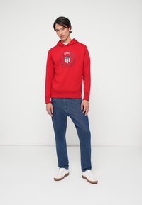 Hoodie rosso con un logo tondo e dettagli grafici sul petto, abbinato a jeans blu larghi e sneakers bianche su uno sfondo neutro.