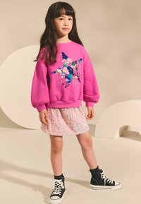 Hanorac roz cu un design din paiete în formă de stea, asortat cu o fustă pastelată strălucitoare și teneși negri înalți. Fundalul prezintă culori deschise.