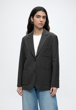Giovane donna con lunghi capelli scuri indossa un blazer grigio scuro sopra una maglietta bianca e jeans azzurri, in piedi davanti a uno sfondo chiaro uniforme.