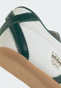 Adidas Tokyo tenisica koja ima bijelu tkaninu gornji dio s zelenim kožnim detaljima, cik-cak šavovima i teksturiranim bež gumom potplatom.