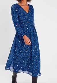 Robe à imprimé floral bleu avec des manches longues, décolleté en V et taille ajustée, comportant des accents dorés et une jupe fluide.