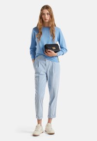 Frau in hellblauem Pullover und Hose mit weißen Sneakern, die eine schwarze Tasche vor einem schlichten weißen Hintergrund hält.