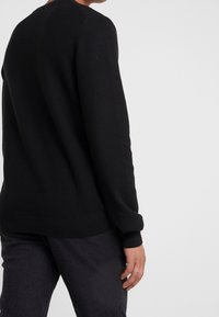 Black knit sweater with textured fabric, crew neck, and ribbed cuffs, worn with dark pants, focusing on the back and sleeve detail.