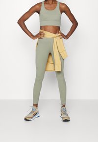 Zelená žebrovaná sportovní podprsenka a odpovídající legíny s žlutým svetrem uvázaným v pase. Bežové a modré sportovní boty dotvářejí outfit.