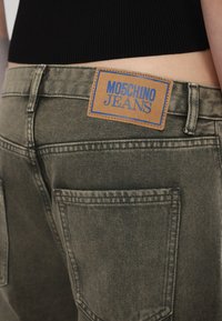 Dunkelolivgrüne Denim-Jeans mit Gesäßtaschen und einem hellbraunen Lederetikett mit blauer Schrift, auf dem "MOSCHINO JEANS" steht.