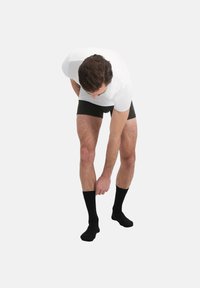 Calzini neri, boxer neri e una maglietta bianca. La persona si piega leggermente, aggiustando il calzino con le dita. Texture della pelle liscia visibile.