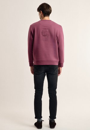 Sudadera burdeos con un diseño texturizado en la parte trasera, cuello redondo, mangas largas y puños acanalados, combinada con jeans oscuros y zapatos marrones.