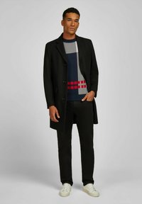 Mann in einem schwarzen Mantel, gemustertem marineblauen Pullover, schwarzen Hosen und weißen Sneakern, der mit einer Hand in der Tasche vor einem grauen Hintergrund steht.