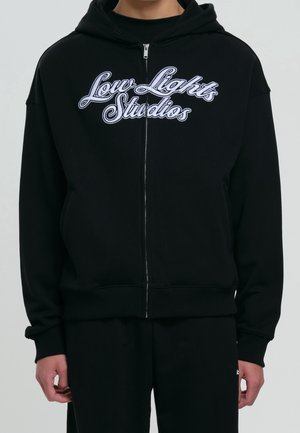 Osoba ubrana w czarną bluzę z zamkiem, z białym napisem „Low Lights Studios” na piersi na jednolitym tle.