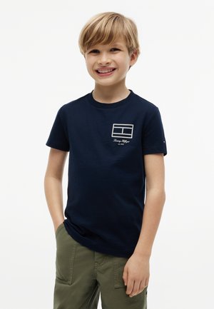 Joven sonriente con cabello rubio que lleva una camiseta azul marino con un pequeño logo blanco y pantalones cargo verde oliva.