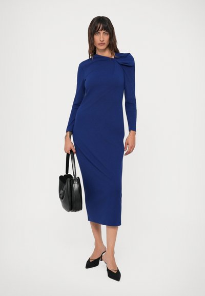 Mulher vestida com um vestido midi de mangas longas em azul royal, com um detalhe assimétrico em forma de laço, segurando uma bolsa preta e usando sapatos de salto pretos e pontiagudos.