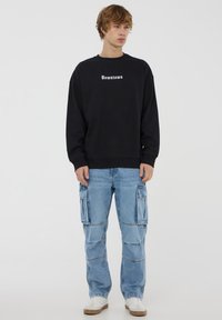 Felpa nera con testo "Downtown", abbinata a jeans cargo azzurri chiari con grandi tasche laterali e sneaker bianche.