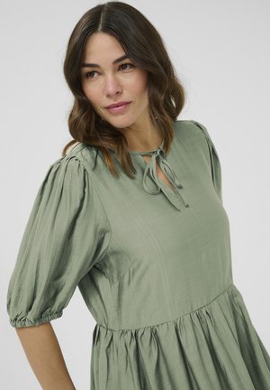 Grøn kjole lavet af tekstureret stof, med puffede ærmer, en snorekrave og en flagrende nederdel med rynkede detaljer.