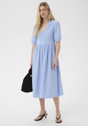 Žena s dlhými blond vlasmi v svetlomodrých midi šatách s nadýchanými rukávmi a čiernymi sandálmi na podpätku, drží čiernu kabelku.