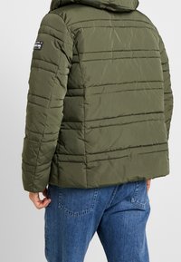 Oliwkowa kurtka puchowa z poziomym pikowaniem, z odpinanym kapturem i minimalnym logo na górnej części rękawa. Noszona z niebieskimi dżinsami.