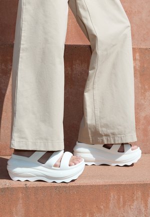 Sandales à plateforme blanches avec une semelle épaisse et ondulée, bout ouvert, et deux brides superposées. Portées avec un pantalon large beige clair.
