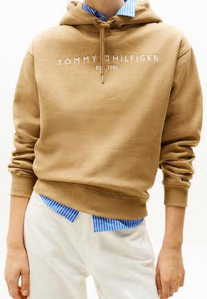 Person iført en beige Tommy Hilfiger hættetrøje over en blåstribet skjorte, kombineret med hvide bukser, hænder afslappet ved siderne.