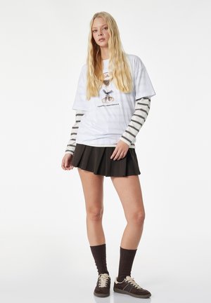 Junge Frau mit langen blonden Haaren, die ein weißes grafisches T-Shirt über gestreiften langen Ärmeln, einen schwarzen plissierten Rock, dunkle Socken und braune Sneaker trägt.