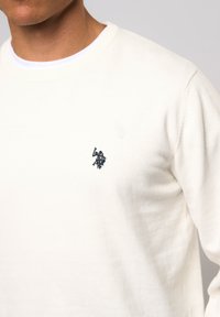 Pull-over en crème clair avec un col ras du cou et des bordures côtelées. Présente un petit logo brodé bleu marine sur la poitrine. Texture douce.