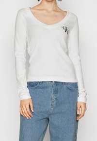 Vit långärmad V-ringad topp i ribbat tyg, med en diskret logotyp på bröstet. Kombinerad med högmidjade jeans.