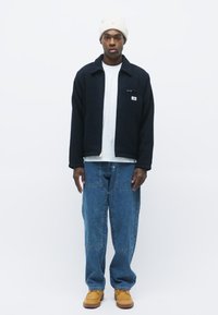Chaqueta de forro polar negra con cremallera sobre una camiseta blanca, jeans de mezclilla azul holgados y botas marrones. El modelo lleva un gorro crema. Diseño simple.
