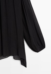 Blouse noire à manches longues bouffantes. En tissu léger froissé. L'ourlet est asymétrique, plus long sur les côtés.