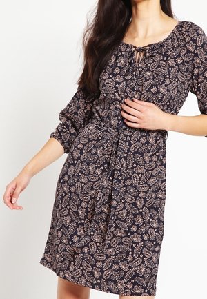 Kvinde iført en knælængde, mørk blomsterkjole med trekvartærmer og snøre i halsen, poserer med den ene hånd på hoften.